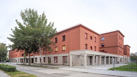 Langes, terrakottafarbenes Gebäude mit profilierter Fassade; Straßenbäume und überdachter Eingang an der Ecke.
