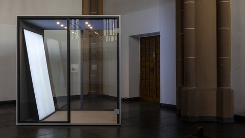 Gläserner Kubus mit beleuchteter Fläche als Kunstinstallation im Innenraum einer Kirche.