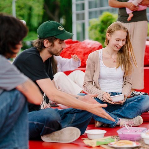 Gruppe junger Menschen sitzt auf roten Sitzkissen im Freien und teilt Speisen bei einem Picknick