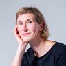 „Porträt einer lächelnden Frau mit kurzen blonden Haaren im schwarzen Oberteil, die ihren Kopf sanft auf die Hand stützt.“