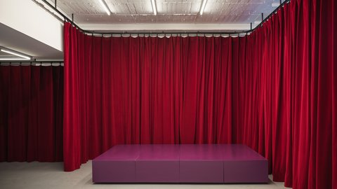 "Ein leerer Raum mit grauem Boden und einer Decke mit sichtbaren Leuchtstoffröhren. Die Wände sind vollständig mit schweren, bodenlangen roten Samtvorhängen bespannt. Mittig im Raum steht ein lila-pinkfarbenes, niedriges, stufenartiges Podest. Die Szene wirkt wie eine leere Bühne oder ein Vorbereitungsraum."  Bild 5: