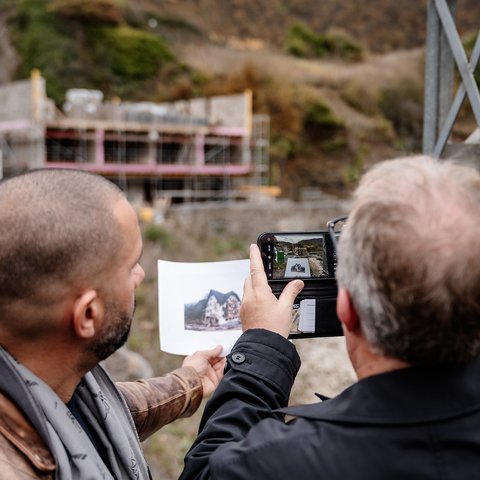 Zwei Männer vor einer Baustelle im Rohbau: Einer hält ein gedrucktes Bild des fertigen Gebäudes, der andere fotografiert dieses mit seinem Smartphone.