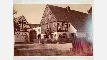 Altes, sepiafarbenes Schwarz-Weiß-Foto auf Fotopapier, das einen ländlichen Dorfplatz zeigt. Links steht ein Fachwerkhaus mit sichtbaren schwarzen Balken. Rechts davon ein größeres Fachwerkhaus mit einem Schieferdach und einer steinernen Veranda mit einem großen Holztor. Im Vordergrund, über die gesamte Breite des Fotos, erstreckt sich ein Holzzaun, der einen unbefestigten Platz begrenzt. Ein Baum steht links vor dem Fachwerkhaus. Das Foto hat leichte Lichtreflexionen auf der Oberfläche.