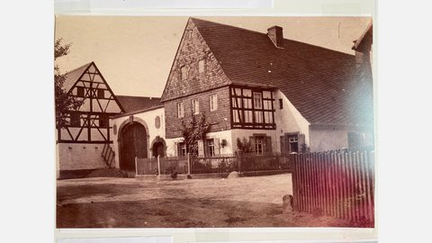 Altes, sepiafarbenes Schwarz-Weiß-Foto auf Fotopapier, das einen ländlichen Dorfplatz zeigt. Links steht ein Fachwerkhaus mit sichtbaren schwarzen Balken. Rechts davon ein größeres Fachwerkhaus mit einem Schieferdach und einer steinernen Veranda mit einem großen Holztor. Im Vordergrund, über die gesamte Breite des Fotos, erstreckt sich ein Holzzaun, der einen unbefestigten Platz begrenzt. Ein Baum steht links vor dem Fachwerkhaus. Das Foto hat leichte Lichtreflexionen auf der Oberfläche.