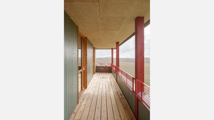 Überdachter Laubengang mit Holzbohlen, grüner Fassade und roten Stützen; Blick über offene Felder. Lang Geradliniger Balkon/Laubengang mit Holzboden, niedriger Brüstung und rotem Netzgeländer. Grüne vertikale Holzverschalung an der Wand, rote Holzstützen und helle Deckenplatten; am Ende kleiner Tisch mit Stuhl, weite Landschaft dahinter.
