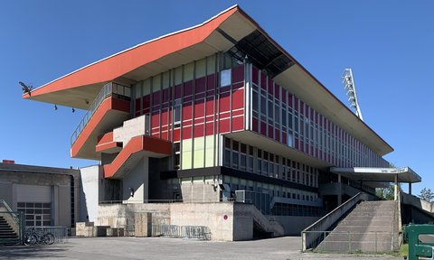 Außenansicht eines älteren Tribünengebäudes mit markantem überstehendem Dach und einer rot-weißen, teils verglasten Fassade bei blauem Himmel.