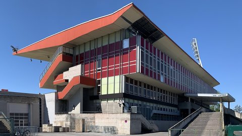 Außenansicht eines älteren Tribünengebäudes mit markantem überstehendem Dach und einer rot-weißen, teils verglasten Fassade bei blauem Himmel.