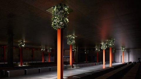 "Dunkler Ausstellungsraum mit einem zentralen Laufsteg. Schlanke, orange beleuchtete Säulen tragen oben dichte Blumenarrangements. Um den Laufsteg stehen einfache Sitzblöcke. Die Beleuchtung hebt die Struktur und Tiefe des Raums hervor."