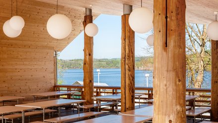 Überdachte Terrasse mit Holztischen auf Rollen und runden Pendelleuchten; Rundholzstützen, Blick auf den See.