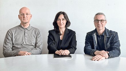 "Porträt von drei Personen, die an einem weißen Tisch vor einer hellen Wand sitzen. Links ein Mann mit Glatze und Brille in grauem Hemd, in der Mitte eine Frau mit dunklen Haaren in schwarzem Blazer, rechts ein Mann mit Brille in Jeansjacke. Alle blicken zur Kamera."