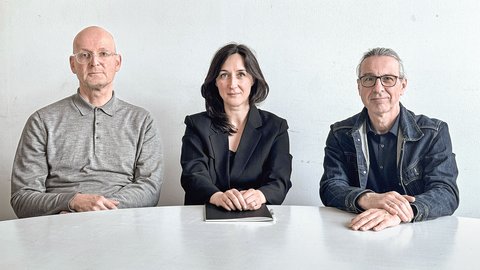 "Porträt von drei Personen, die an einem weißen Tisch vor einer hellen Wand sitzen. Links ein Mann mit Glatze und Brille in grauem Hemd, in der Mitte eine Frau mit dunklen Haaren in schwarzem Blazer, rechts ein Mann mit Brille in Jeansjacke. Alle blicken zur Kamera."
