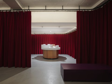 "Eine Innenansicht eines Raumes, der komplett von bodenlangen, roten Samtvorhängen umgeben ist. Durch einen Vorhangspalt im Vordergrund blickt man auf einen runden Holztisch mit weißer Platte, auf dem ein architektonisches Modell steht. Im Vordergrund, vor dem Vorhang, befindet sich ein niedriges, lila-pinkfarbenes Möbelstück."