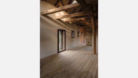 "Großer heller Innenraum mit minimalistischem Design. Weiße Wände und Decke. Massive Holzbalken an der Decke spannen sich über den Raum. Links eine große Glastür mit schwarzem Rahmen. Mehrere kleinere Fenster mit schwarzen Rahmen in der Wand. Heller Holzfußboden aus breiten Dielen."