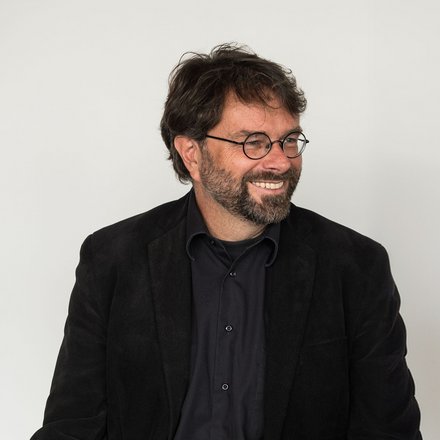 "Porträtfoto eines Mannes mittleren Alters vor hellem, neutralem Hintergrund. Er trägt eine schwarze Brille mit runder Fassung, einen schwarzen Blazer und ein dunkelblauens Hemd. Er hat dunkelbraunes, lockiges Haar und einen dunklen Bart. Der Mann lächelt und schaut nach links aus dem Bild."
