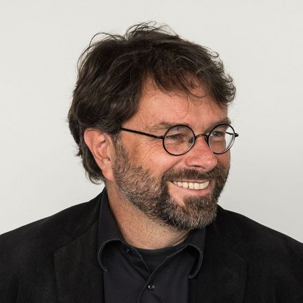 "Porträtfoto eines Mannes mittleren Alters vor hellem, neutralem Hintergrund. Er trägt eine schwarze Brille mit runder Fassung, einen schwarzen Blazer und ein dunkelblauens Hemd. Er hat dunkelbraunes, lockiges Haar und einen dunklen Bart. Der Mann lächelt und schaut nach links aus dem Bild."