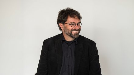 "Porträtfoto eines Mannes mittleren Alters vor hellem, neutralem Hintergrund. Er trägt eine schwarze Brille mit runder Fassung, einen schwarzen Blazer und ein dunkelblauens Hemd. Er hat dunkelbraunes, lockiges Haar und einen dunklen Bart. Der Mann lächelt und schaut nach links aus dem Bild."