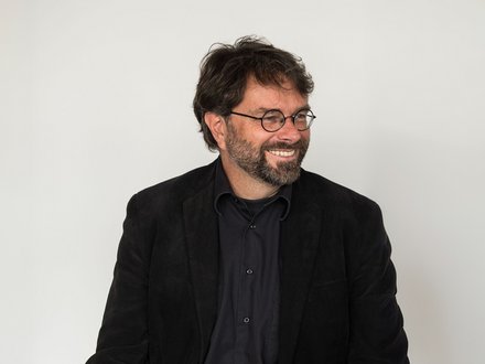 "Porträtfoto eines Mannes mittleren Alters vor hellem, neutralem Hintergrund. Er trägt eine schwarze Brille mit runder Fassung, einen schwarzen Blazer und ein dunkelblauens Hemd. Er hat dunkelbraunes, lockiges Haar und einen dunklen Bart. Der Mann lächelt und schaut nach links aus dem Bild."