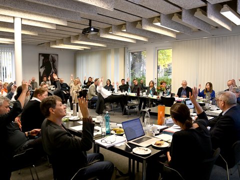 Menschen in einem Besprechungsraum sitzen an Tischen in U-Form und heben mehrheitlich die rechte Hand