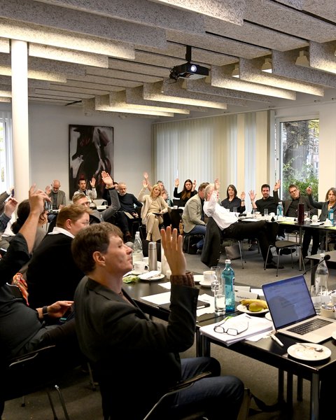 Menschen in einem Besprechungsraum sitzen an Tischen in U-Form und heben mehrheitlich die rechte Hand