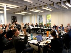 Menschen in einem Besprechungsraum sitzen an Tischen in U-Form und heben mehrheitlich die rechte Hand