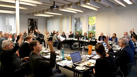 Menschen in einem Besprechungsraum sitzen an Tischen in U-Form und heben mehrheitlich die rechte Hand