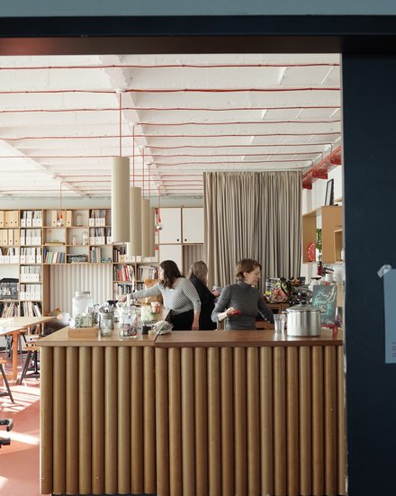 Innenraum der Cafeteria der Leibniz Universität Hannover mit modernem Design. Im Vordergrund eine Theke mit vertikalen Holzlamellen, dahinter drei Mitarbeiterinnen. Der Raum zeichnet sich durch eine hohe, helle Decke mit roten Designelementen aus. Sichtbar sind Bücherregale an den Wänden, ein Stehtisch, grüne Pflanzen und eine beige Gardine. Der Boden ist terracottafarben. Links neben dem Eingang sind Plakate und rechts ein Informationsdisplay zu sehen."