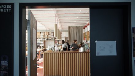 Innenraum der Cafeteria der Leibniz Universität Hannover mit modernem Design. Im Vordergrund eine Theke mit vertikalen Holzlamellen, dahinter drei Mitarbeiterinnen. Der Raum zeichnet sich durch eine hohe, helle Decke mit roten Designelementen aus. Sichtbar sind Bücherregale an den Wänden, ein Stehtisch, grüne Pflanzen und eine beige Gardine. Der Boden ist terracottafarben. Links neben dem Eingang sind Plakate und rechts ein Informationsdisplay zu sehen."