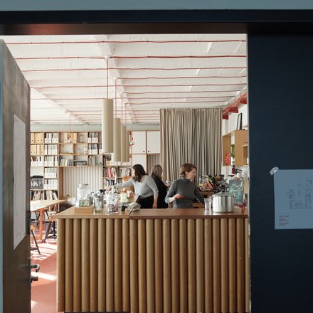 Innenraum der Cafeteria der Leibniz Universität Hannover mit modernem Design. Im Vordergrund eine Theke mit vertikalen Holzlamellen, dahinter drei Mitarbeiterinnen. Der Raum zeichnet sich durch eine hohe, helle Decke mit roten Designelementen aus. Sichtbar sind Bücherregale an den Wänden, ein Stehtisch, grüne Pflanzen und eine beige Gardine. Der Boden ist terracottafarben. Links neben dem Eingang sind Plakate und rechts ein Informationsdisplay zu sehen."