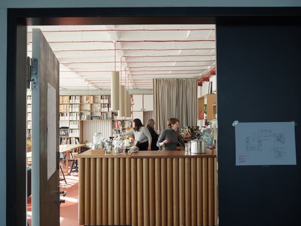 Innenraum der Cafeteria der Leibniz Universität Hannover mit modernem Design. Im Vordergrund eine Theke mit vertikalen Holzlamellen, dahinter drei Mitarbeiterinnen. Der Raum zeichnet sich durch eine hohe, helle Decke mit roten Designelementen aus. Sichtbar sind Bücherregale an den Wänden, ein Stehtisch, grüne Pflanzen und eine beige Gardine. Der Boden ist terracottafarben. Links neben dem Eingang sind Plakate und rechts ein Informationsdisplay zu sehen."