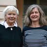 "Ein Porträtfoto von zwei älteren Frauen, die nebeneinander stehen und in die Kamera lächeln. Die linke Frau hat kurzes, weißes Haar und trägt einen schwarzen Pullover mit einer weißen Bluse darunter und einer Brosche. Die rechte Frau hat längeres, lockiges, grau-braunes Haar und trägt ein blau-weiß gestreiftes Langarmshirt."