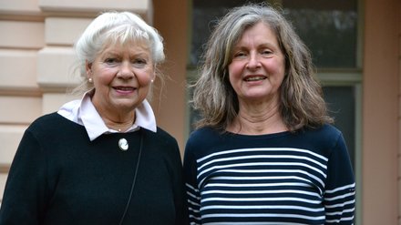 "Ein Porträtfoto von zwei älteren Frauen, die nebeneinander stehen und in die Kamera lächeln. Die linke Frau hat kurzes, weißes Haar und trägt einen schwarzen Pullover mit einer weißen Bluse darunter und einer Brosche. Die rechte Frau hat längeres, lockiges, grau-braunes Haar und trägt ein blau-weiß gestreiftes Langarmshirt."