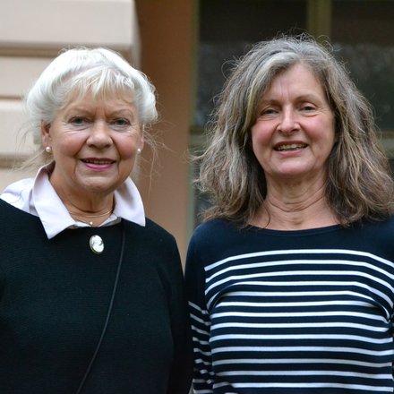 "Ein Porträtfoto von zwei älteren Frauen, die nebeneinander stehen und in die Kamera lächeln. Die linke Frau hat kurzes, weißes Haar und trägt einen schwarzen Pullover mit einer weißen Bluse darunter und einer Brosche. Die rechte Frau hat längeres, lockiges, grau-braunes Haar und trägt ein blau-weiß gestreiftes Langarmshirt."