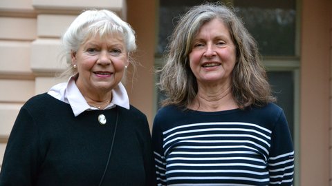 "Ein Porträtfoto von zwei älteren Frauen, die nebeneinander stehen und in die Kamera lächeln. Die linke Frau hat kurzes, weißes Haar und trägt einen schwarzen Pullover mit einer weißen Bluse darunter und einer Brosche. Die rechte Frau hat längeres, lockiges, grau-braunes Haar und trägt ein blau-weiß gestreiftes Langarmshirt."