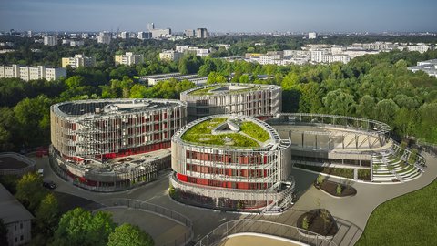 Luftaufnahme eines modernen Gebäudekomplexes mit drei runden, begrünten Dächern umgeben von Bäumen und Stadt im Hintergrund