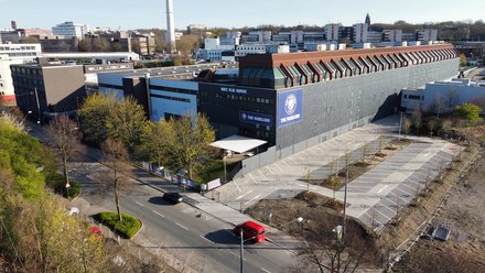 Schräg-Luftaufnahme eines langen, grauen Industriegebäudes mit rotem Sheddach. An der Fassade Logos „THE PADELERS“ und der Schriftzug „MEET. PLAY. REPEAT.“ Rechts ein neuer, fast leerer Parkplatz mit jungen Bäumen; vorne eine Straße mit rotem Kleinbus und schwarzem Auto, im Hintergrund Bürogebäude und ein hoher Schornstein.