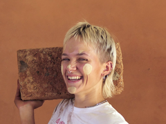 Lächelnde Person mit kurzen blonden Haaren, Lehmflecken im Gesicht und auf dem T-Shirt, die einen großen Lehmbaustein auf der Schulter trägt.