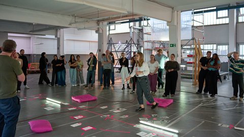 Gruppe von Menschen steht in einem großen Raum um auf dem Boden ausgelegte Karten und pinkfarbene Kissen