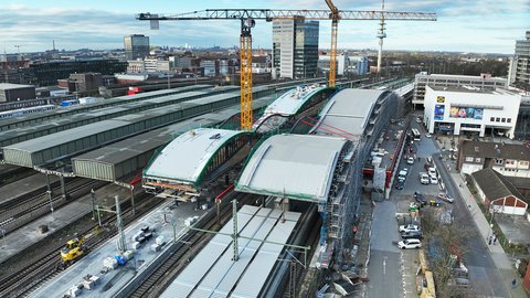 Bahnhof im Umbau (Luftaufnahme)  Eine Luftaufnahme zeigt einen großen Bahnhof mit mehreren Gleisen und Bahnsteigen. Über den Gleisen befinden sich große, gebogene Bahnsteigdächer im Bauzustand, teilweise eingerüstet. Zwei hohe gelbe Kräne überspannen die Anlage. Rechts verläuft eine Straße mit Autos, links sind weitere Bahnanlagen und Industriegebäude zu sehen. Das Bild zeigt den Umbau und die Modernisierung eines zentralen Verkehrsknotens. Es steht für Infrastrukturentwicklung, Mobilität und die bauliche Anpassung öffentlicher Verkehrsräume.