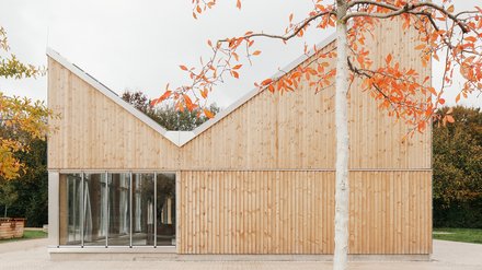 Front des Holzpavillons mit V‑förmiger Dachsilhouette; junger Baum mit orangefarbenen Blättern im Vordergrund auf gepflastertem Platz.