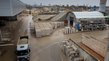 Übersicht eines Baustoffwerks mit Kieshaufen, gestapelten Betonblöcken, Lagerhallen und einem Lkw mit Schüttgut.