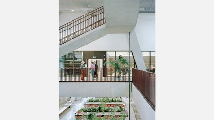 Mehrstöckiges Treppenhaus mit hellem Innenraum und braunem Geländer. Im unteren Bereich befinden sich Sitzgelegenheiten umgeben von Pflanzen. Im mittleren Bereich befinden sich drei Personen und dahinter offene Türen und Glasfronten.