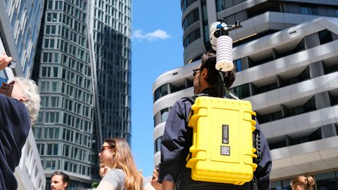 Nahaufnahme von hinten auf einen Mann mit einem auffälligen gelben Rucksack mit Messgeräten inmitten moderner Hochhäuser.