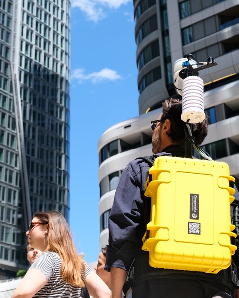 Nahaufnahme von hinten auf einen Mann mit einem auffälligen gelben Rucksack mit Messgeräten inmitten moderner Hochhäuser.