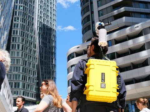 Nahaufnahme von hinten auf einen Mann mit einem auffälligen gelben Rucksack mit Messgeräten inmitten moderner Hochhäuser.