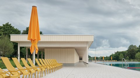 "Modernes Freibad fotografiert von der Liegewiese aus. Im Vordergrund eine lange Reihe gelber Liegestühle auf beiger Betonplattform. Zentral steht ein großer, zusammengefalteter orangefarbener Sonnenschirm auf dunklem Ständer. Dahinter erstreckt sich ein weiß-beiges Flachdachgebäude mit offener Konstruktion und beigen Lamellenwänden. Auf der rechten Seite ist ein türkisblauer Pool mit Metallleiter, rotem Abgrenzungsseil und weiteren orangefarbenen Schirmen sichtbar. Links und rechts grüne Vegetation und Bäume. Der Himmel ist bewölkt und grau, mit Wolkenformationen im oberen Bildbereich."