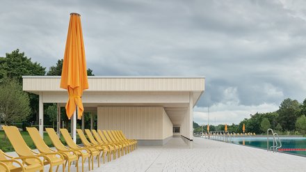 "Modernes Freibad fotografiert von der Liegewiese aus. Im Vordergrund eine lange Reihe gelber Liegestühle auf beiger Betonplattform. Zentral steht ein großer, zusammengefalteter orangefarbener Sonnenschirm auf dunklem Ständer. Dahinter erstreckt sich ein weiß-beiges Flachdachgebäude mit offener Konstruktion und beigen Lamellenwänden. Auf der rechten Seite ist ein türkisblauer Pool mit Metallleiter, rotem Abgrenzungsseil und weiteren orangefarbenen Schirmen sichtbar. Links und rechts grüne Vegetation und Bäume. Der Himmel ist bewölkt und grau, mit Wolkenformationen im oberen Bildbereich."