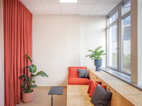 "Gemütlicher Ruheraum mit Fensterfront. Links großer korallrot-farbener Vorhang zum Abdunkeln. Holzbank-Fensterbank mit orangenen Kissen und grauem Kissen. Rechts große Fenster mit Blick nach draußen. Eine grüne Topfpflanze steht auf der Bank. Kleine schwarze Ablage. Weiße Wände, Holzboden, warme natürliche Beleuchtung."
