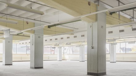 Leerer Hallenraum mit Betonstützen  Ein großer, heller Raum mit regelmäßig angeordneten Betonstützen. Die Decke ist niedrig, mit sichtbaren Leuchten und technischen Installationen. Der Raum ist vollständig leer. Das Bild zeigt die räumliche Qualität des Bestands: Offenheit, Wiederholbarkeit und strukturelle Klarheit als Grundlage für neue Programme.