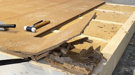 Geöffnete Ecke eines Holzrahmen‑Bauelements: Holzfaser‑Dämmung sichtbar, gelöste Platten; Handwerkzeuge liegen oben, Werkhalle im Hintergrund.