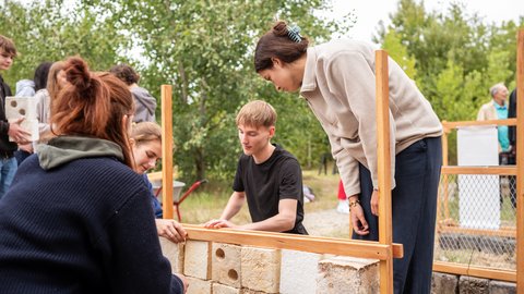 „Mehrere Personen arbeiten im Freien gemeinsam an einer niedrigen Mauer, indem sie helle Bausteine in ein Holzgerüst schichten.“